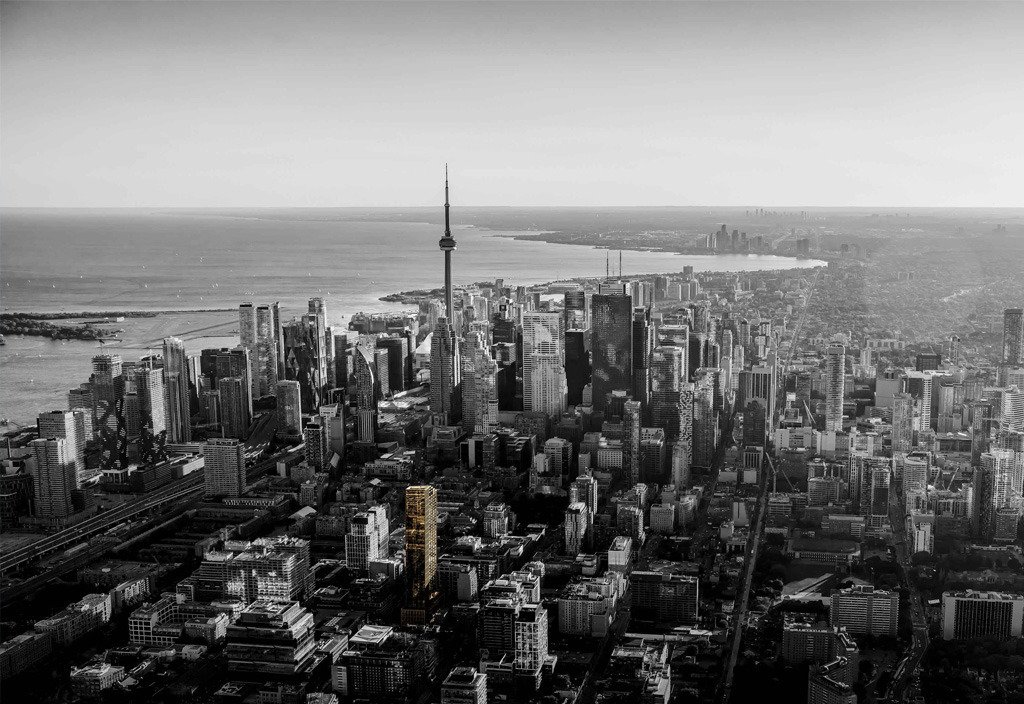 Aerial view of a sprawling cityscape with a prominent tower at its center. The city is bordered by a large body of water on the left, and new condos in Brampton stand out among the monochrome buildings, highlighted by one in yellow.