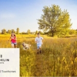A young man and woman run joyfully with a golden retriever through a sunlit field of tall grass, with trees in the background under a clear blue sky. In the bottom left corner, a Brooklin Heights townhomes logo and text that reads "Brooklin Heights | Townhomes" are displayed.