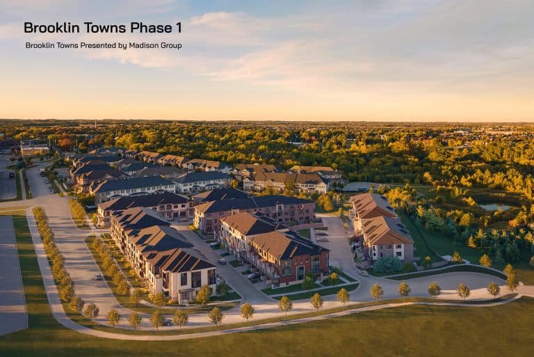 Aerial view of a suburban residential development called "Brooklin Towns Phase 1" by Madison Group. The image shows rows of townhouses with driveways and green spaces, set against a background of trees and a partly cloudy sky at sunset. Brooklin Towns promises tranquil living amidst natural beauty.