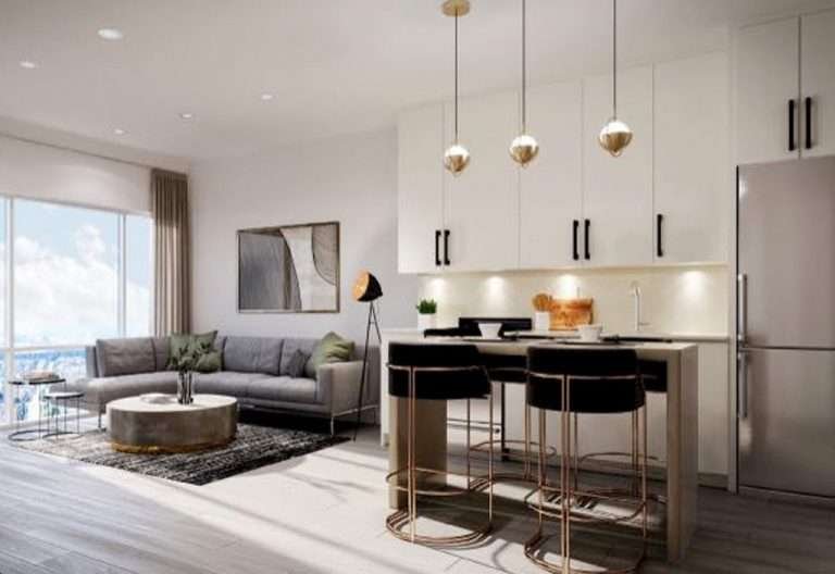 Modern open-concept living room and kitchen area with sleek white cabinetry, stainless steel appliances, and pendant lights. The living area has a gray sectional sofa, round coffee table, floor lamp, and large window with a view. New condos in GTA feature stylish rugs and wall art to complete the look.