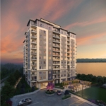 A multi-story apartment building stands against a picturesque sunset sky. The modern structure, showcasing new condos in Brampton, features balconies on each floor. A car is parked in the foreground, and there's a landscaped entrance area with seating. A body of water is visible in the background.