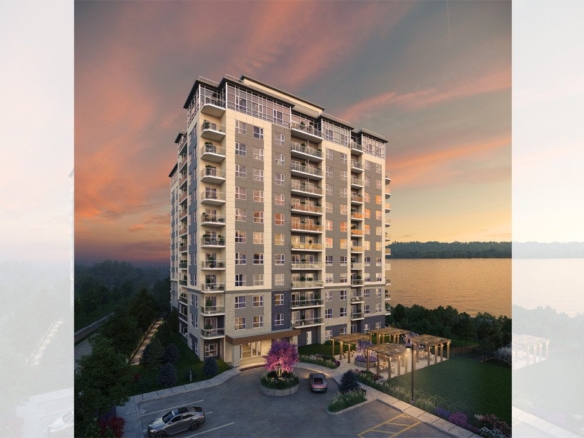 A multi-story apartment building stands against a picturesque sunset sky. The modern structure, showcasing new condos in Brampton, features balconies on each floor. A car is parked in the foreground, and there's a landscaped entrance area with seating. A body of water is visible in the background.