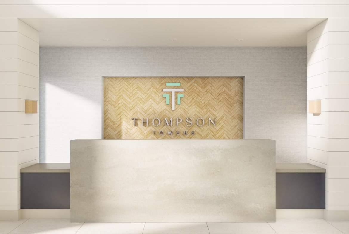 A modern reception desk with a light gray counter stands in front of a wall featuring a wood herringbone pattern. Above the desk, the text "THOMPSON TOWERS" is displayed alongside a stylized "TT" logo. The area, well-lit with neutral tones and minimalist design, highlights the appeal of new condos in GTA.