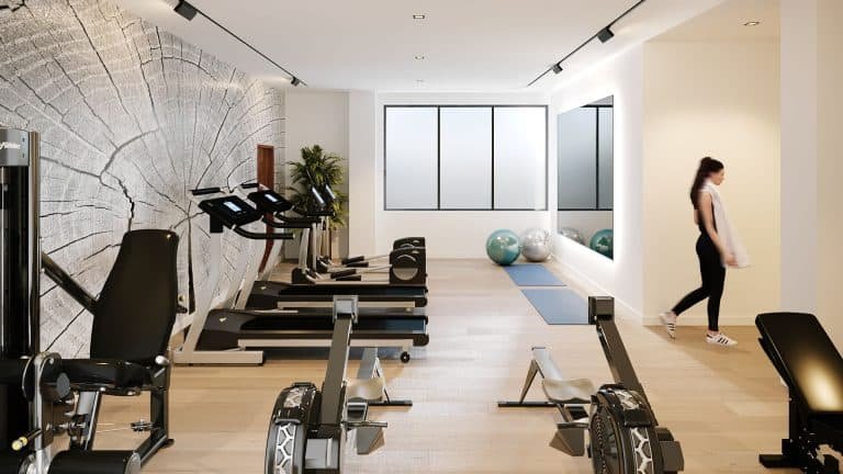 A modern gym at Motto Condos features treadmills, rowing machines, and exercise bikes lined up against a wall with a large tree ring mural. A woman stands near exercise balls and yoga mats, with bright windows bringing in natural light in the background—an ideal real estate investment for fitness enthusiasts.
