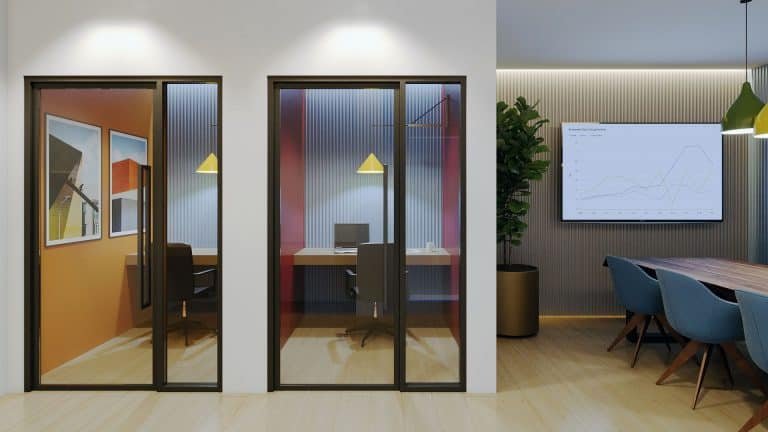A modern office space at Motto Condos features two glass-walled private rooms with single desks and chairs. The main area boasts a conference table surrounded by blue chairs, a large plant, and a wall-mounted screen displaying charts. Artwork adorns the walls of the private rooms, enhancing the sleek design.