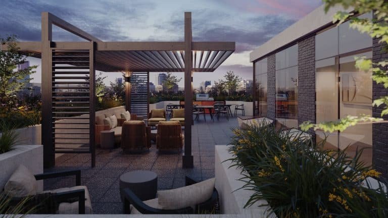 A modern rooftop terrace at dusk at Motto Condos features cushioned seating, a wooden pergola, a BBQ area, and lush potted plants. The city skyline is visible in the background under a colorful sky. The space is illuminated by subtle ambient lighting, creating an inviting atmosphere.
