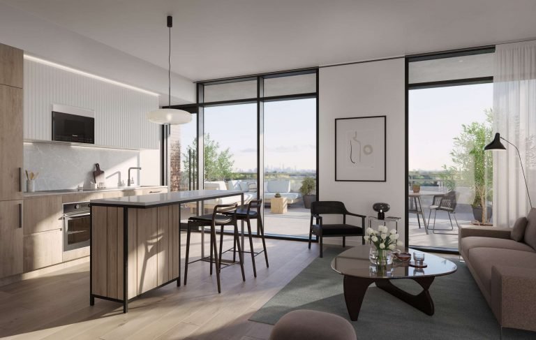 A modern, open-plan living area at Six99 Condos features a kitchen with wooden cabinetry, an island with seating, and a cozy lounge area. Large glass doors lead to a spacious outdoor balcony with views of the greenery and cityscape in the distance.