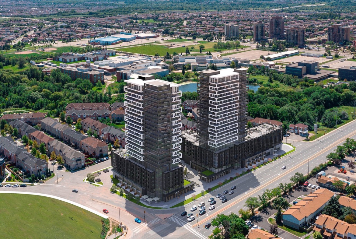 Aerial view of two tall residential towers under New condos construction beside a busy highway, surrounded by a suburban landscape with houses, smaller buildings, and patches of greenery.