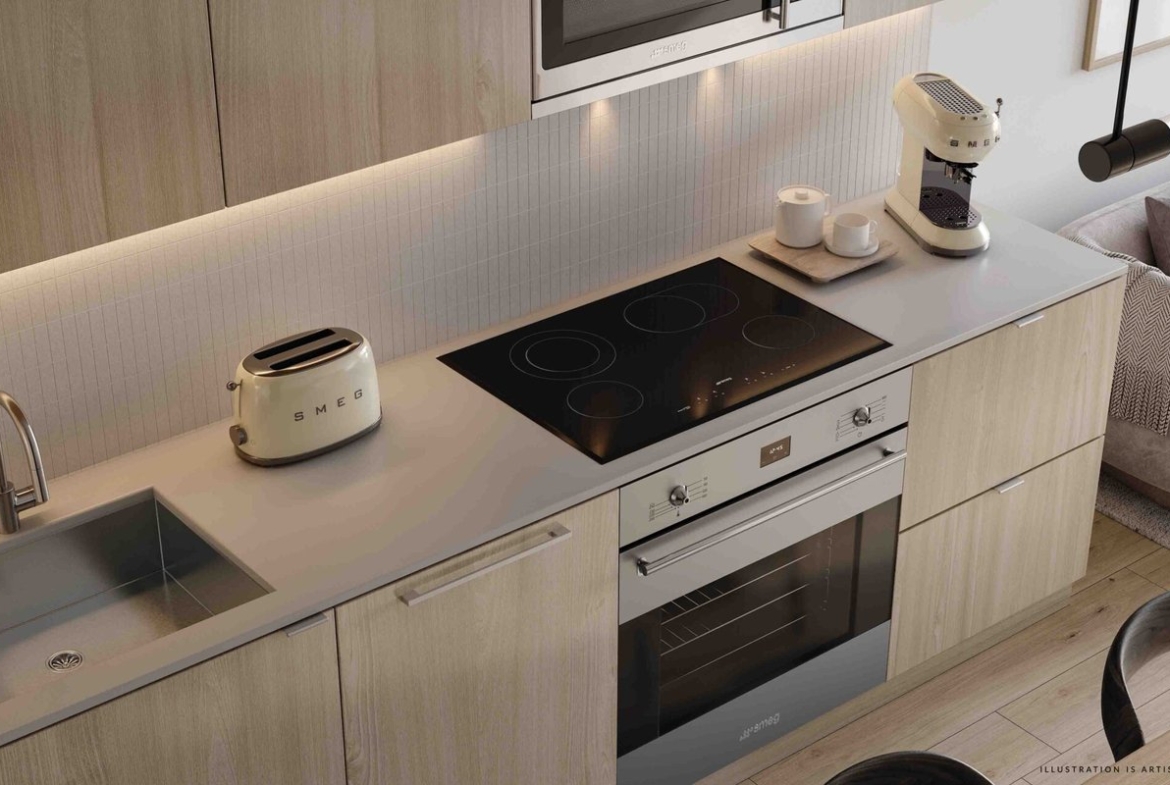 Modern kitchen interior featuring light wood cabinets, a sleek induction stove, built-in oven, and small appliances in a pre-construction home. A cozy seating area with a gray sofa is visible adjacent