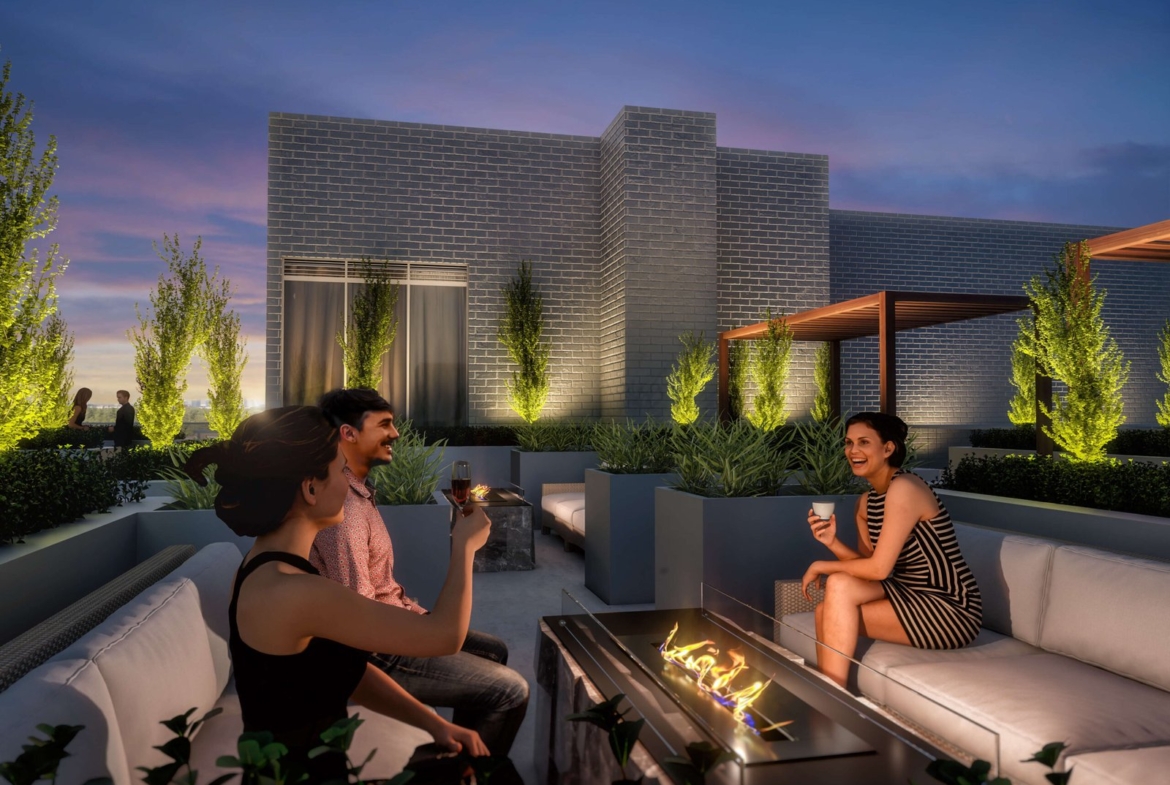 Three people are relaxing on a rooftop lounge in the evening, sipping drinks around a fire pit. The modern setting features brick buildings, lush greenery, and ambient lighting, perfectly complementing the dusky sky with its blend of purple and blue hues—a serene spot in a city known for its new homes.
