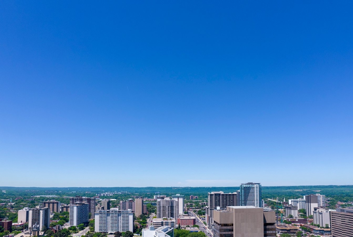 A cityscape featuring numerous high-rise buildings and skyscrapers, set against a clear blue sky. The horizon reveals green, hilly terrain in the background, blending urban and natural landscapes with new condos in GTA offering fresh perspectives on modern living.