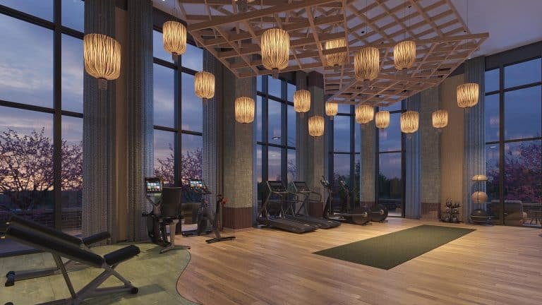 Modern gym in New Homes in GTA with large windows overlooking a sunset, featuring treadmills, elliptical machines, and hanging wooden light fixtures.