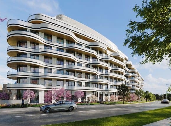 Modern wavy-designed apartment building with spacious balconies, surrounded by pink flowering trees, a lush green lawn, and a clear blue sky, situated beside a quiet street. Ideal for those looking for Pre