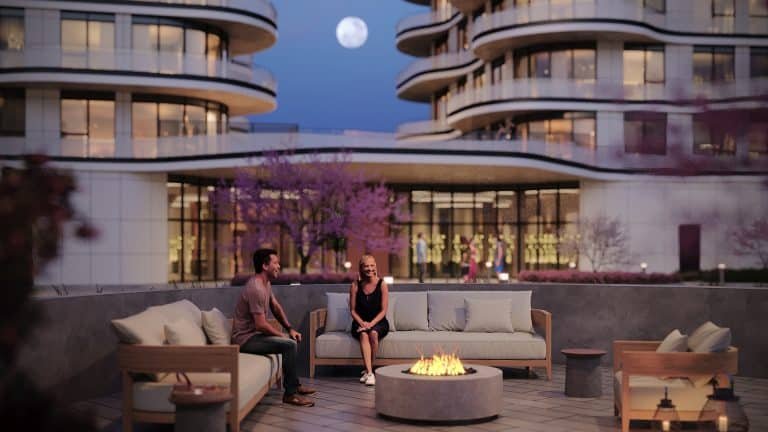A man and woman sit around a lit fire pit on a rooftop patio with modern buildings and a full moon in the background. The scene is set during twilight, with warm ambient lighting from the surrounding buildings and blooming lily trees adding a touch of color to their serene crosstown escape.