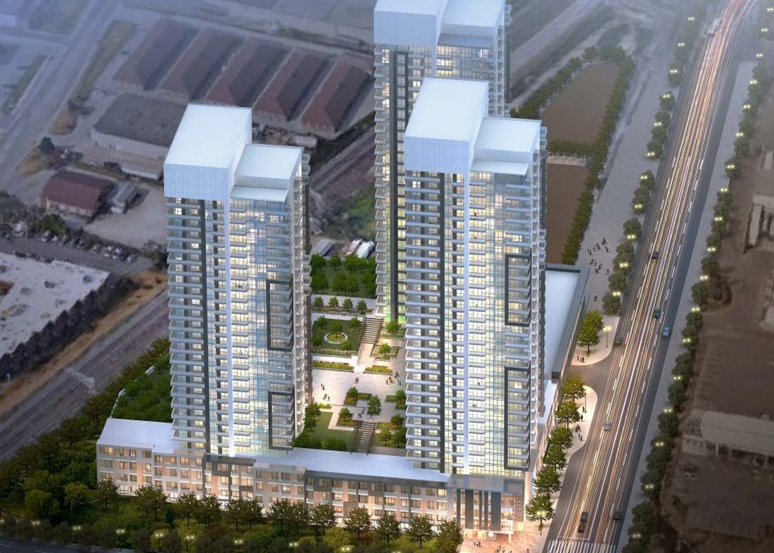 Aerial view of a modern, high-rise residential complex featuring three tall buildings with illuminated windows. The towers are connected by a green rooftop with landscaped gardens. These new condos in GTA are situated by a well-lit, bustling road lined with trees.