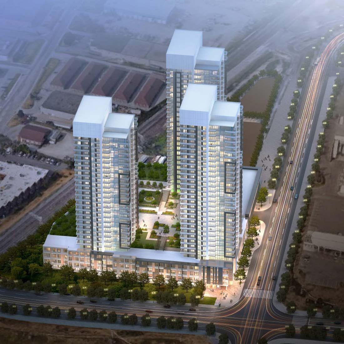 Aerial view of a modern, high-rise residential complex featuring three tall buildings with illuminated windows. The towers are connected by a green rooftop with landscaped gardens. These new condos in GTA are situated by a well-lit, bustling road lined with trees.