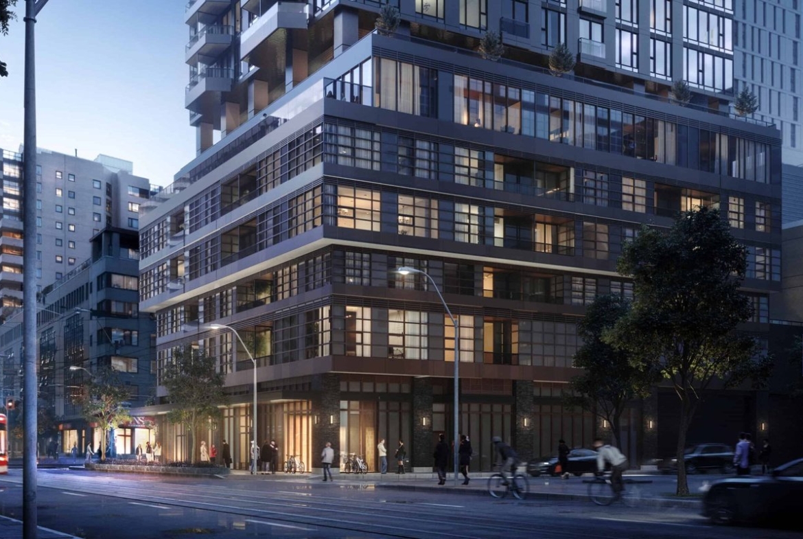 A street view captures a modern city corner at dusk, highlighting a multi-story building with glass windows aglow. Pedestrians stroll along the sidewalk as vehicles and a streetcar navigate the road. Trees line the area, complementing the urban landscape that boasts new condos in GTA.