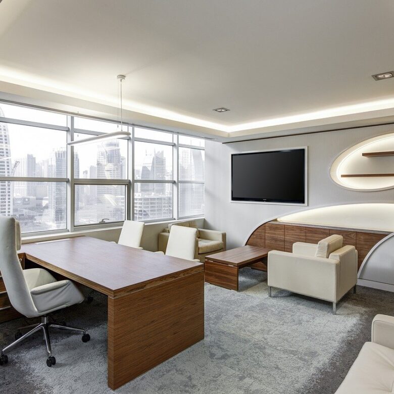 Modern office with wooden desk, white swivel chair, and cream-colored seating area. Large windows offer a city skyline view, reminiscent of the sleek condos in the Dufferin Region. Walls are white with minimalist shelving and a wall-mounted TV. Natural light fills the space.
