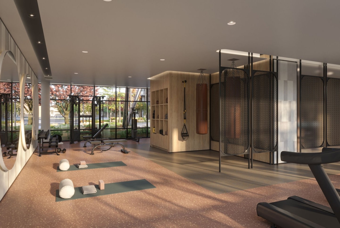 A modern gym interior at Mile & Creek Condos boasts large windows allowing natural light to flood in. Features include yoga mats, exercise balls, a treadmill, a bench, and round mirrors. Outside, trees with pink flowers create a serene view through the windows.