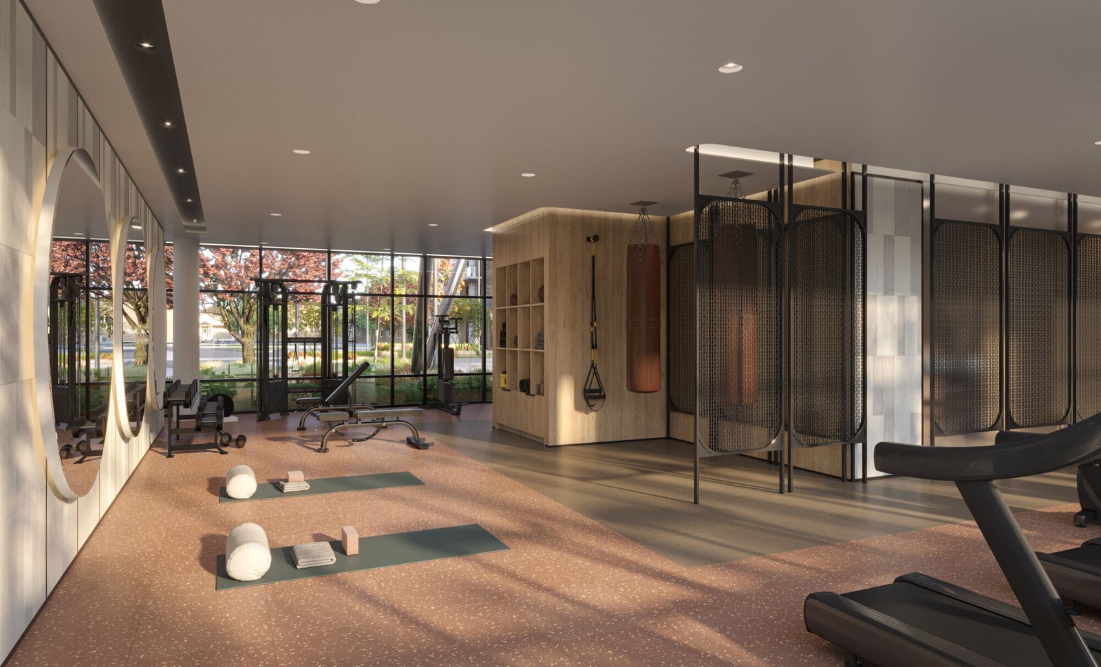 A modern gym interior at Mile & Creek Condos boasts large windows allowing natural light to flood in. Features include yoga mats, exercise balls, a treadmill, a bench, and round mirrors. Outside, trees with pink flowers create a serene view through the windows.