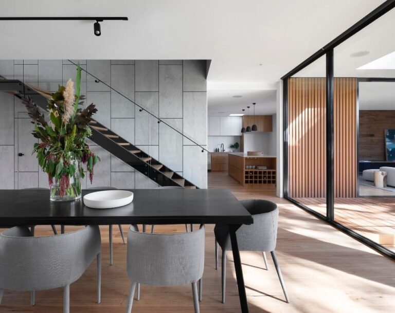 This modern open-plan condo boasts a chic dining area with a sleek black table and gray chairs, complemented by a vibrant floral arrangement. The inviting space includes a staircase, kitchen, and sliding glass doors that open to a sunlit patio—ideal for those seeking new homes in Brampton.