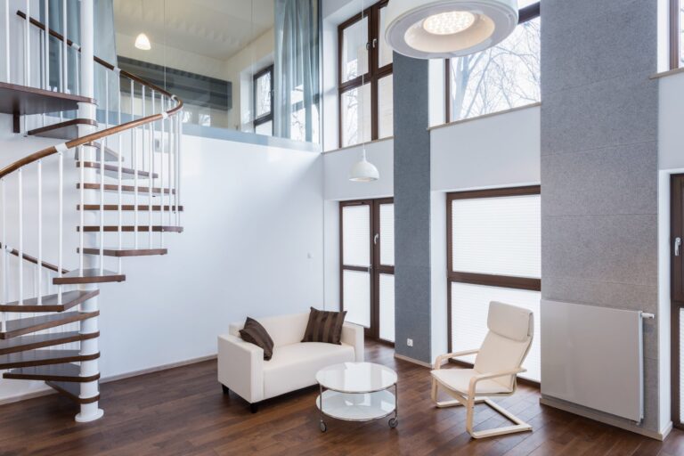 A modern living room with high ceilings, featuring a white sofa, glass coffee table, and beige armchair. A spiral staircase leads to the upper floor. Large windows bring in natural light. This iconic space by New Homes by Gagan Bector boasts wooden flooring and minimalistic decor throughout.
