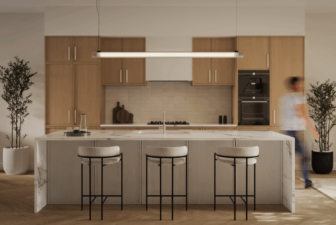 A modern kitchen in a new condo with a marble island and three cushioned barstools. Light wood cabinets complement the linear pendant light above. Stainless steel appliances and minimal decor, including plants, complete the space while a person appears slightly blurred in motion.
