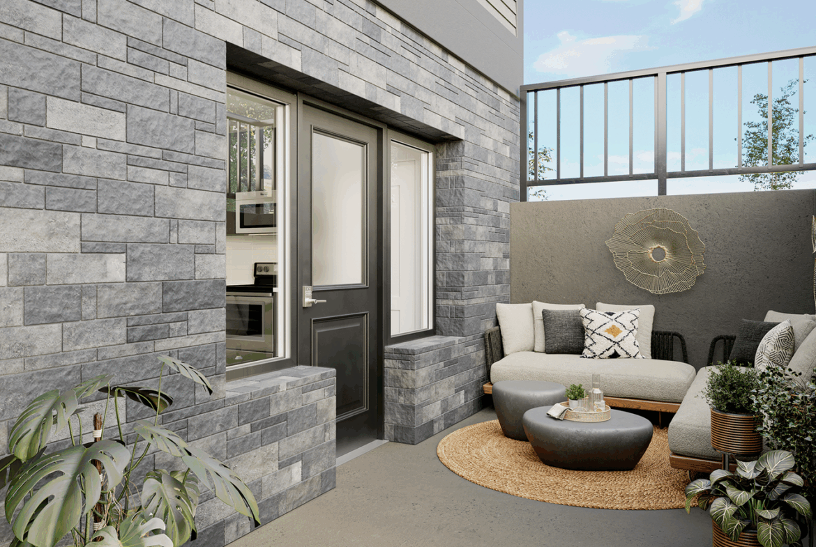 A cozy outdoor patio with a gray brick wall, a beige sectional couch adorned with patterned pillows, and a modern touch from the round woven rug and small table. The area, featuring potted plants and a tall railing, offers a tranquil and stylish Marquis setting.