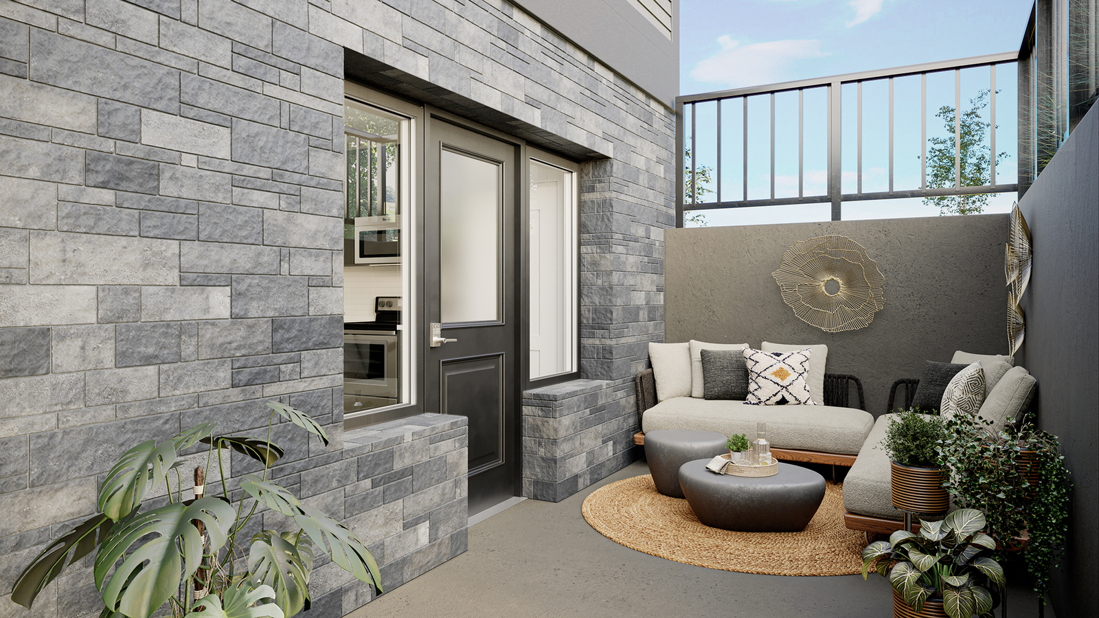 A cozy outdoor patio with a gray brick wall, a beige sectional couch adorned with patterned pillows, and a modern touch from the round woven rug and small table. The area, featuring potted plants and a tall railing, offers a tranquil and stylish Marquis setting.