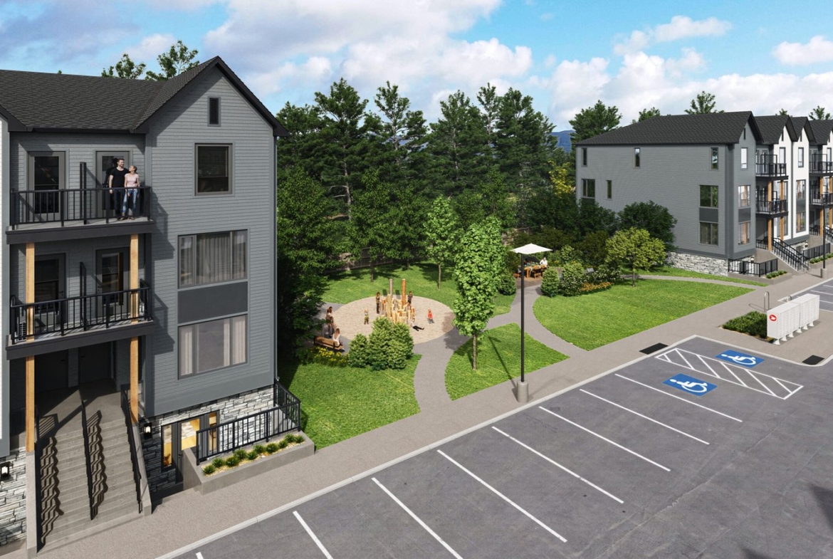 A birds-eye view of a modern apartment complex featuring multiple three-story buildings showcases new homes. A small park with a playground is situated between the buildings. In the foreground, a parking lot includes marked accessible spaces, while trees border this inviting area.