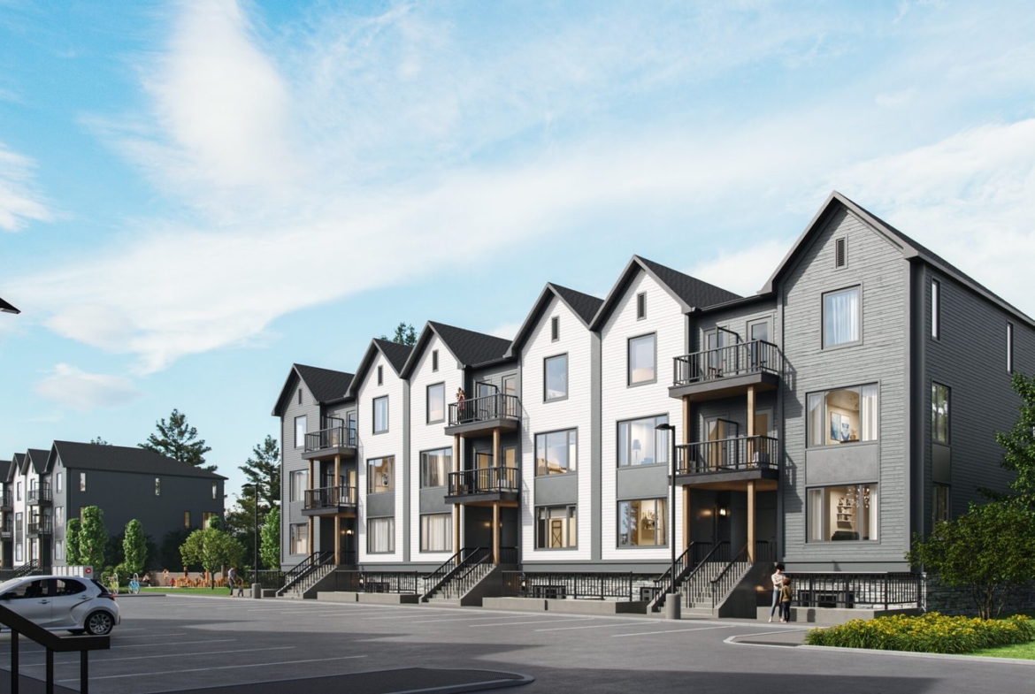 A modern residential complex showcases new homes arranged in two rows of identical, multi-story townhouses. The buildings feature gray and white exteriors with gabled roofs and small balconies. A spacious parking lot sits in the foreground, surrounded by streetlights and a few trees.