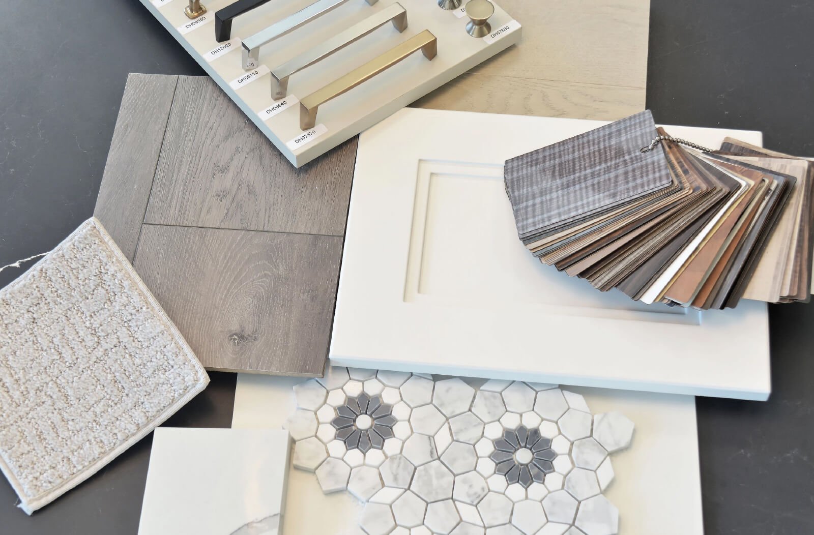 Explore a collection of interior design samples for new homes in Brampton, featuring metallic cabinet handles, wooden flooring, hexagonal marble tiles, and a textured carpet swatch. Discover a variety of colored fabric swatches and a white cabinet door displayed elegantly on a dark surface.