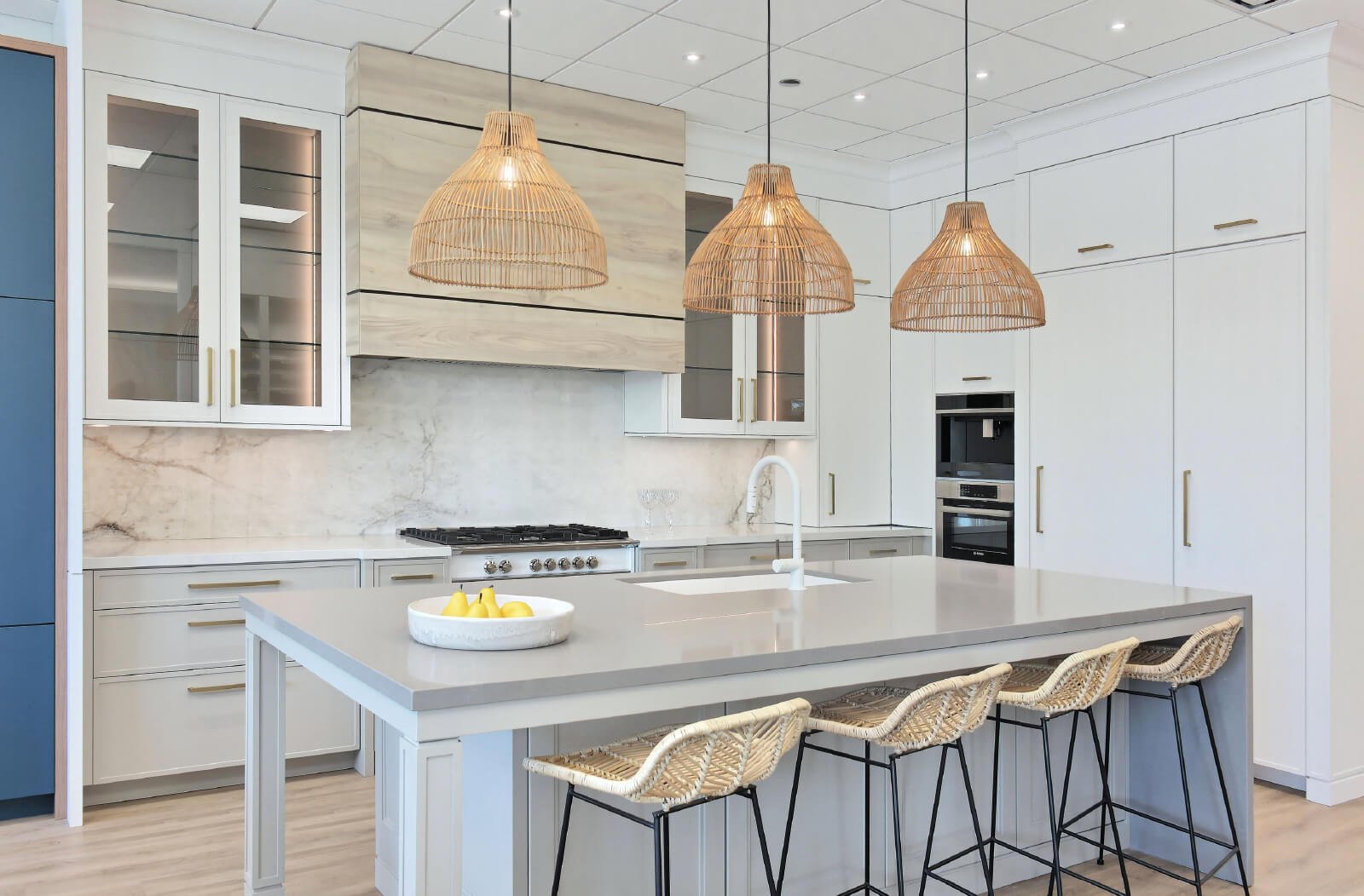 Modern kitchen with a large gray island, featuring three rattan pendant lights and four wicker stools. White cabinets and a marble backsplash create a stunning backdrop. On the island countertop sits a bowl of bananas, perfect for those exploring new homes or pre-construction options in the GTA.