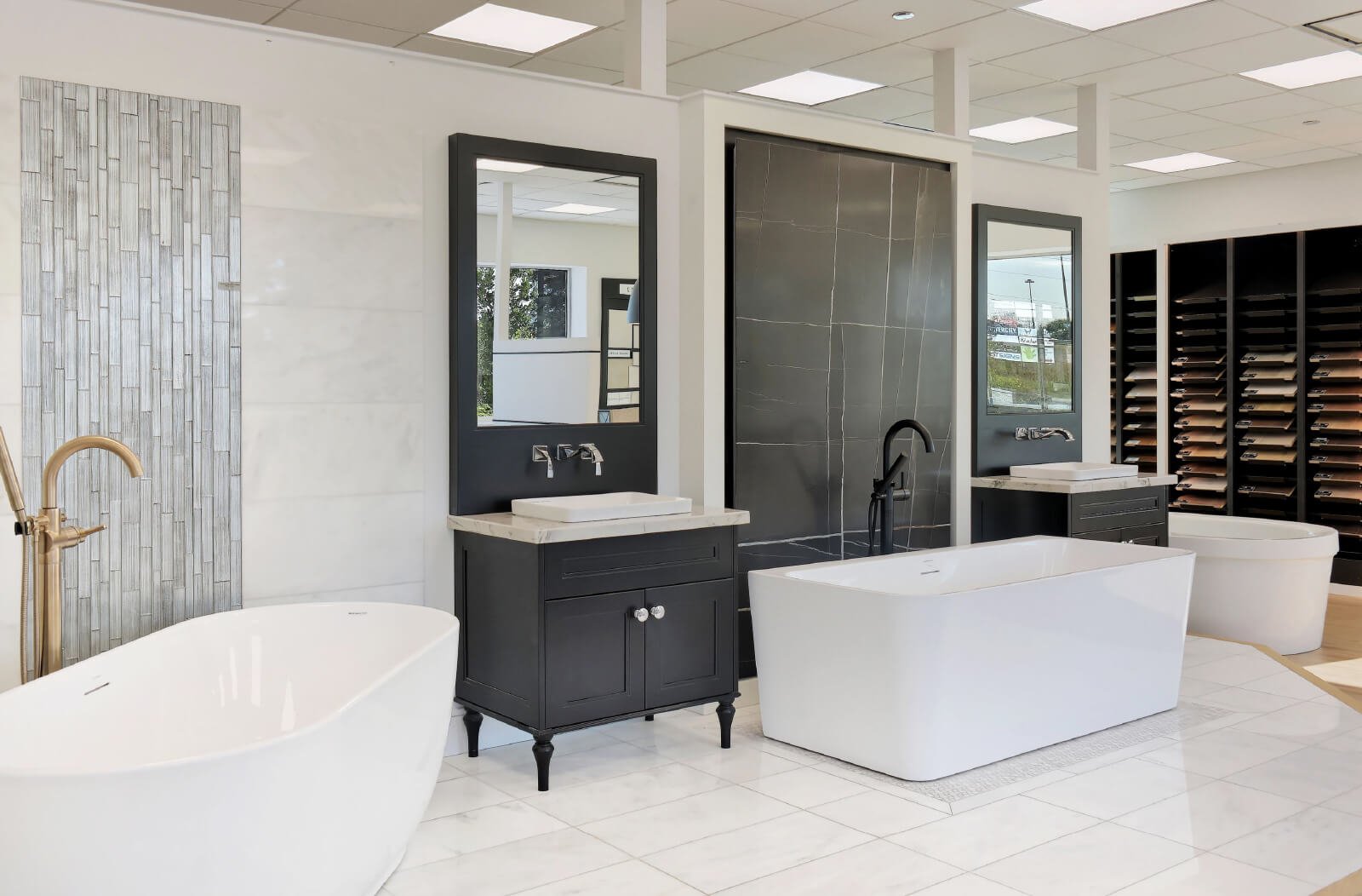 A modern bathroom showroom by Royal Pine Homes in Mayfield Village features an elegant design with freestanding and rectangular tubs, black vanities paired with mirrors, chrome faucets, and a wall display of tile samples.