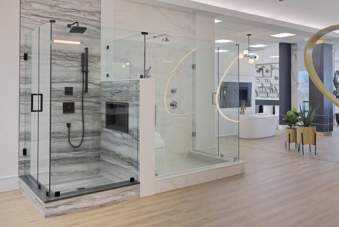 Discover a modern bathroom showroom, perfect for new homes, featuring a glass-enclosed shower with gray and white marble walls and black fixtures. Adjacent is a curved glass section. In the background, contemporary fixtures accompany a white bathtub on elegant wooden flooring.