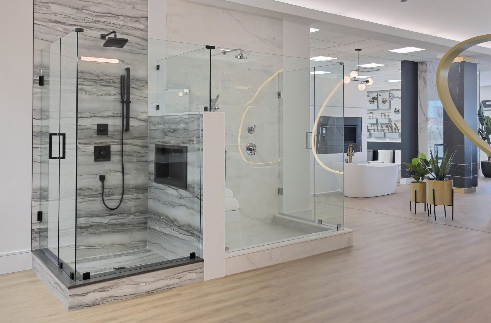 Discover a modern bathroom showroom, perfect for new homes, featuring a glass-enclosed shower with gray and white marble walls and black fixtures. Adjacent is a curved glass section. In the background, contemporary fixtures accompany a white bathtub on elegant wooden flooring.