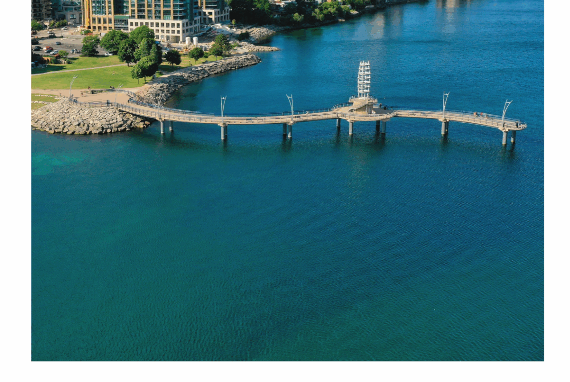 An aerial view of a coastal city with a long pier extending into a blue lake. The foreground showcases the pier and surrounding water, while residential and commercial buildings line the shore. New homes dot the skyline under a sky adorned with clouds.