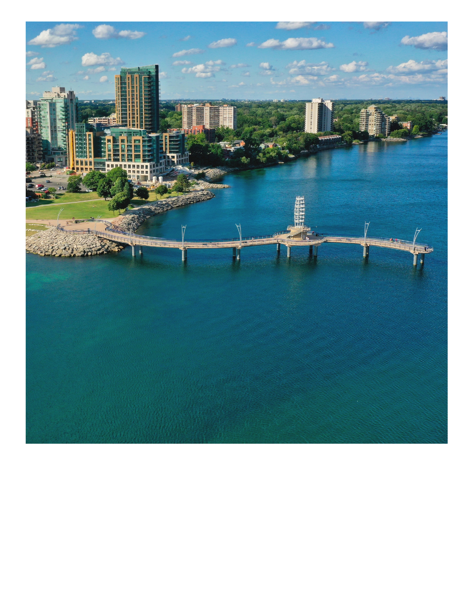 An aerial view of a coastal city with a long pier extending into a blue lake. The foreground showcases the pier and surrounding water, while residential and commercial buildings line the shore. New homes dot the skyline under a sky adorned with clouds.