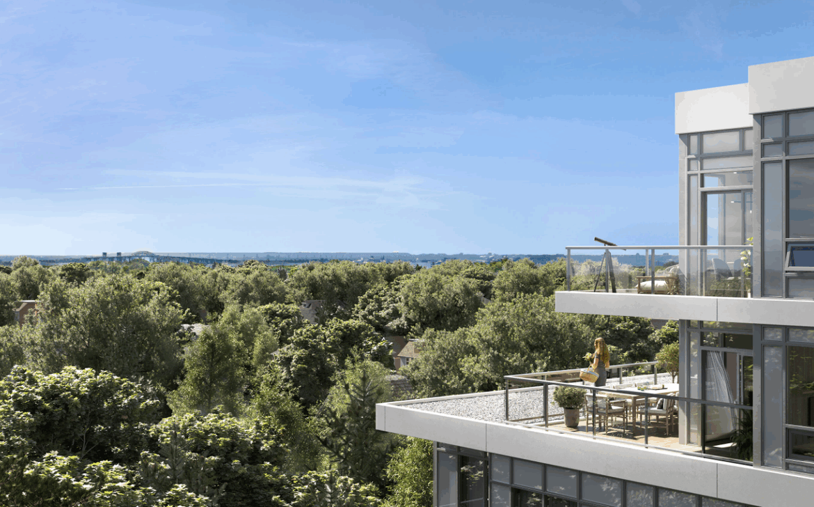 A modern, multi-story building with large glass windows and balconies overlooks a lush green forest. A person stands on a balcony enjoying the view under a clear blue sky, epitomizing the appeal of new homes in Brampton for those craving a scenic escape.