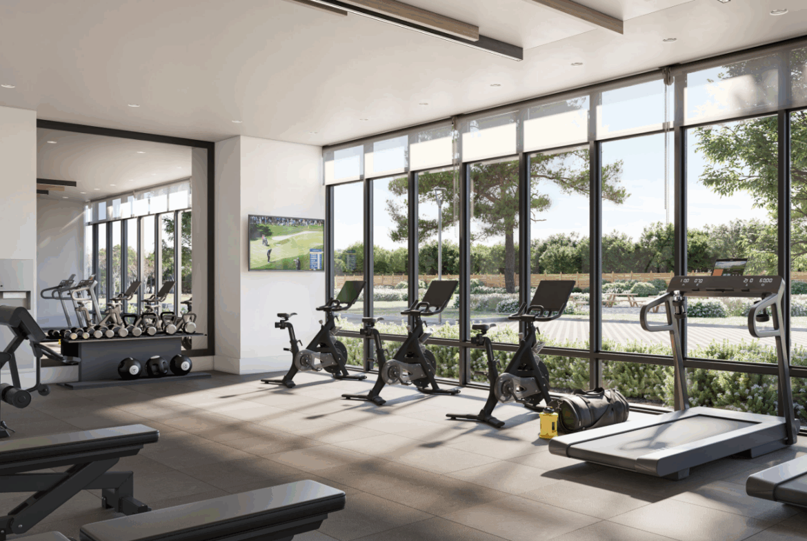 A modern gym with large windows offers a leafy view and includes exercise bikes, treadmills, and weightlifting equipment. Perfect for residents of new homes in Toronto, a TV displays sports while natural light fills the room, highlighting a clean and organized space.