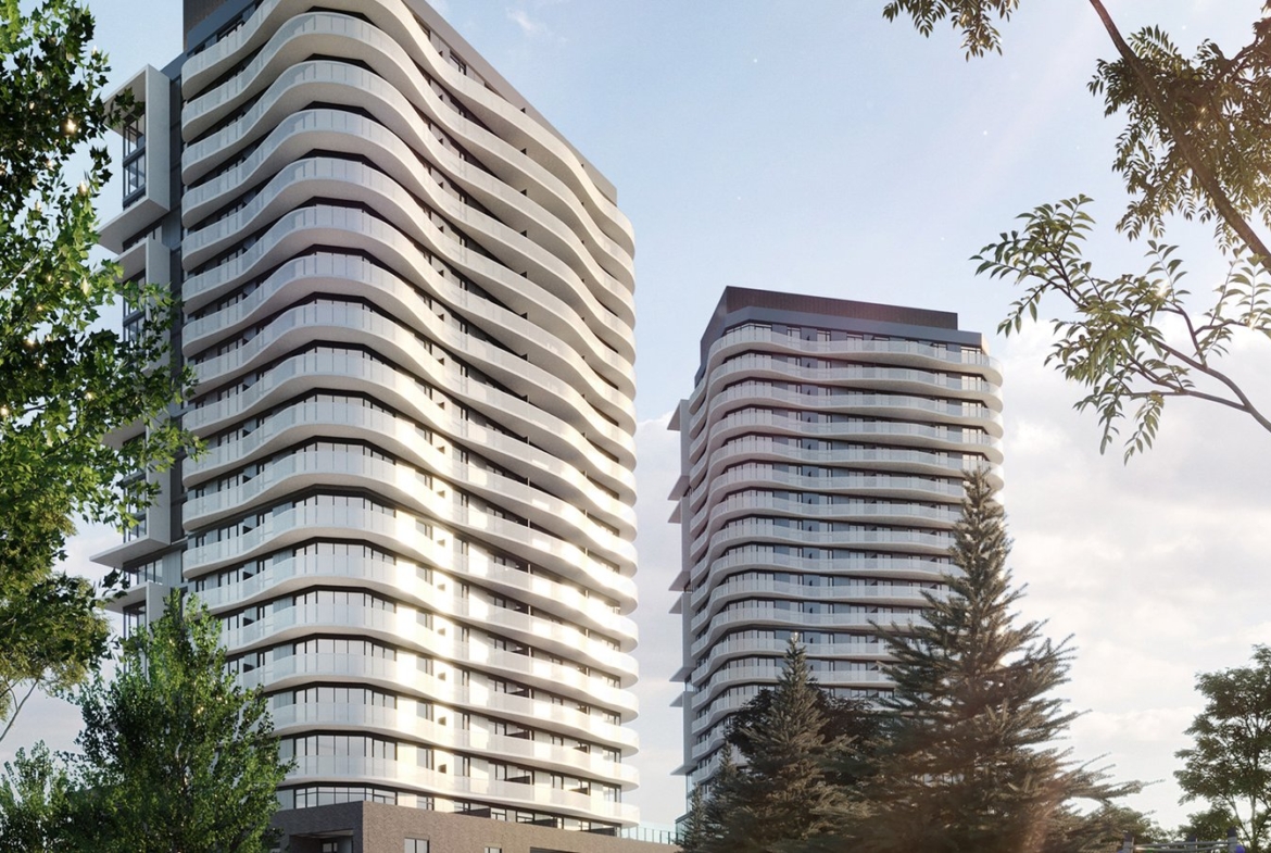 Two modern high-rise buildings with curved balconies stand under a clear sky. The structures, surrounded by trees, add a touch of greenery to this urban setting. Sunlight casts soft shadows on the facades of these new condos in Brampton, offering an impressive blend of nature and architecture.