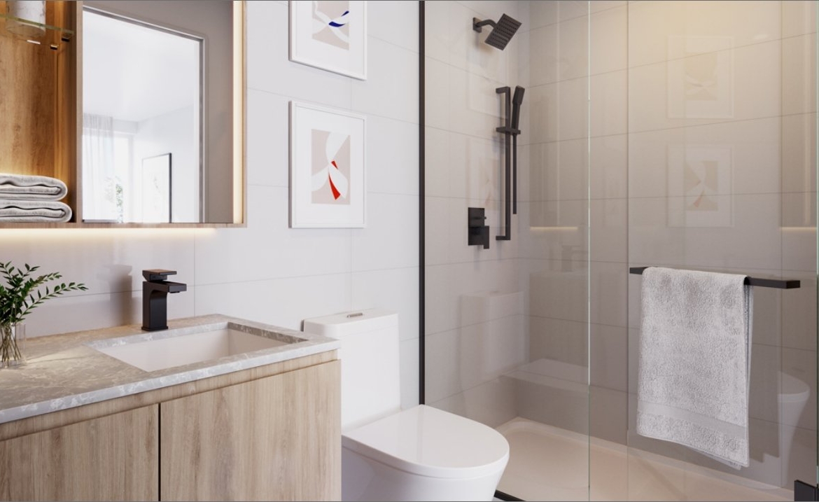 This modern bathroom in the new condos in GTA features a wooden vanity, white countertop, and sleek black fixtures. A large mirror and framed art adorn the wall, while a glass shower door holds a crisp white towel. Natural light floods through a window, enhancing the clean and minimalist design.