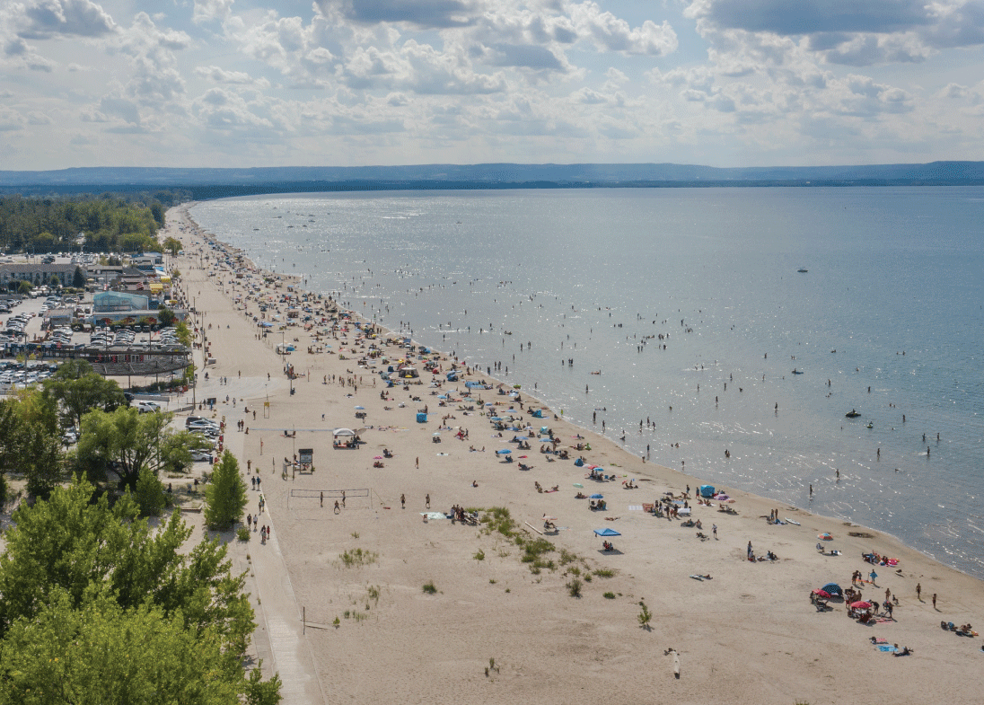 Aerial view of a large, sandy beach with people relaxing and swimming. The beach is dotted with umbrellas and towels under a partly cloudy sky. The ocean stretches to the horizon, while lush greenery borders the shore, reminiscent of serene escapes near new homes in Toronto.
