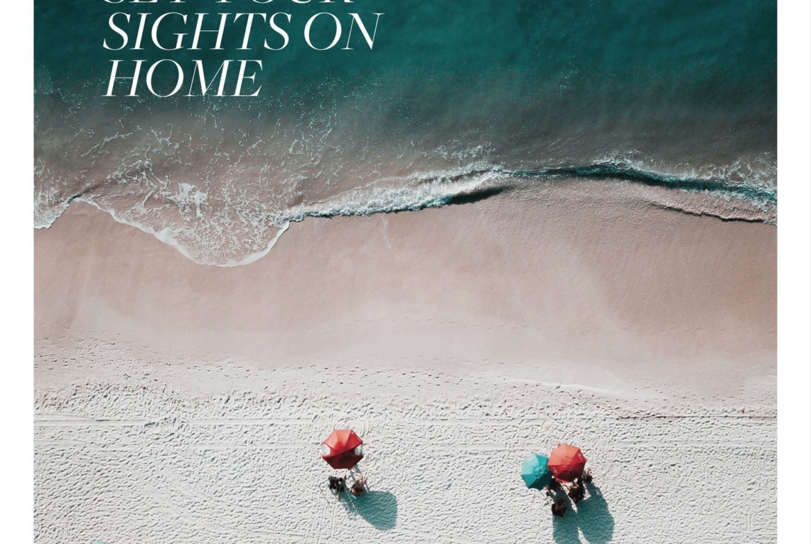 An aerial view of a sandy beach with three people under red and green umbrellas near the water's edge. Text on the image reads, "Set Your Sights on Home." Waves gently lap the shore, inviting you to dream of new homes in Toronto.