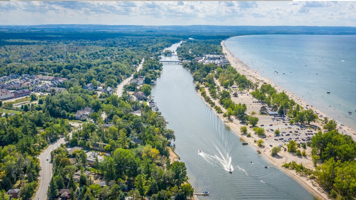 Aerial view of a coastal town with a river running through it, lined with trees and houses. A sandy beach with parked cars and recreational boats on the water is visible. On the horizon, a lush landscape stretches under a partly cloudy sky, hinting at new homes emerging in this picturesque setting.