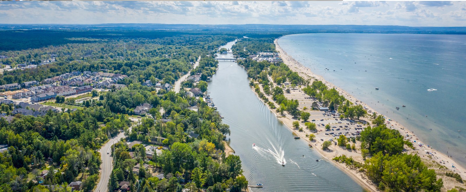Aerial view of a coastal town with a river running through it, lined with trees and houses. A sandy beach with parked cars and recreational boats on the water is visible. On the horizon, a lush landscape stretches under a partly cloudy sky, hinting at new homes emerging in this picturesque setting.