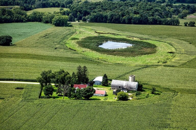 Aerial view of a rural landscape with a farm surrounded by lush green fields, reminiscent of living in Stouffville. The farm features a red-roofed building, barn, and silo. A circular pond graces the fields, bordered by trees and vegetation, offering idyllic new homes in Stouffville settings.
