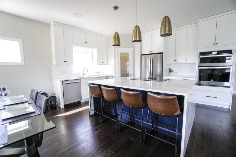 This modern kitchen in the new homes in Shelburne features a large island, brown leather bar stools, white cabinets, and stainless steel appliances accented by pendant lights. A dining table with chairs is visible on the left, while a window bathes the space in natural light over dark wooden flooring.