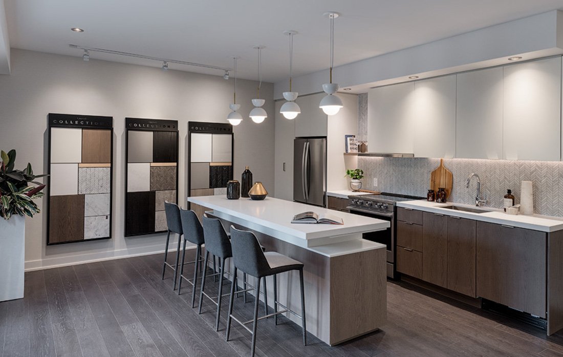 Discover a modern kitchen showroom designed for pre-construction homes, featuring a central island, bar stools, and pendant lights. Sleek countertops and cabinetry complement the wall displays of materials and color samples. Neutral tones and contemporary elements create a sophisticated atmosphere.