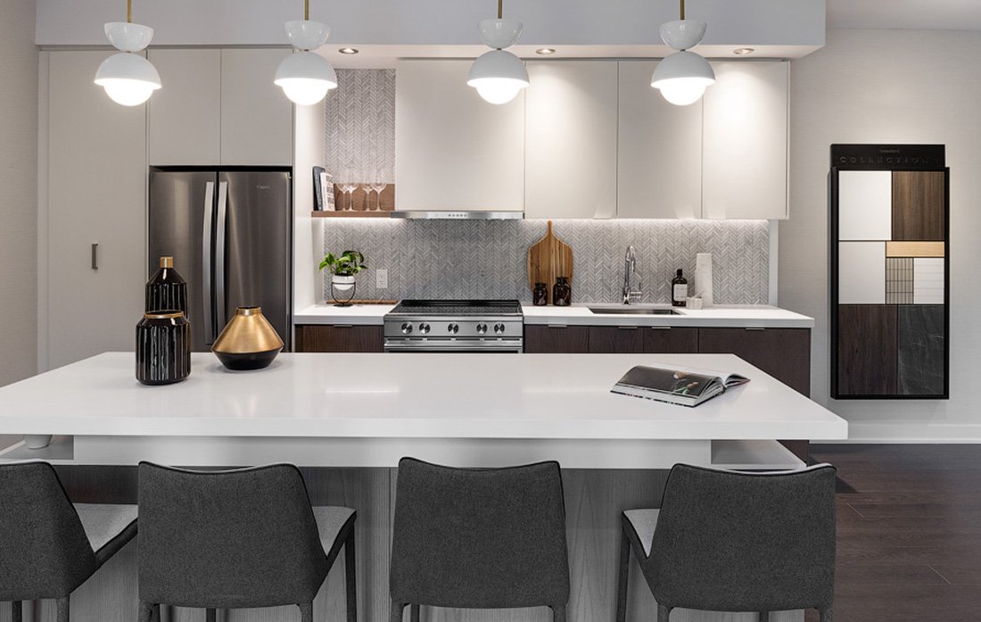 Discover the allure of a modern kitchen in new condos in Brampton, featuring a large white island, gray chairs, and stainless steel appliances. White cabinets pair with decorative vases and art pieces to complement the textured backsplash. Hanging lights elegantly illuminate the sleek space.